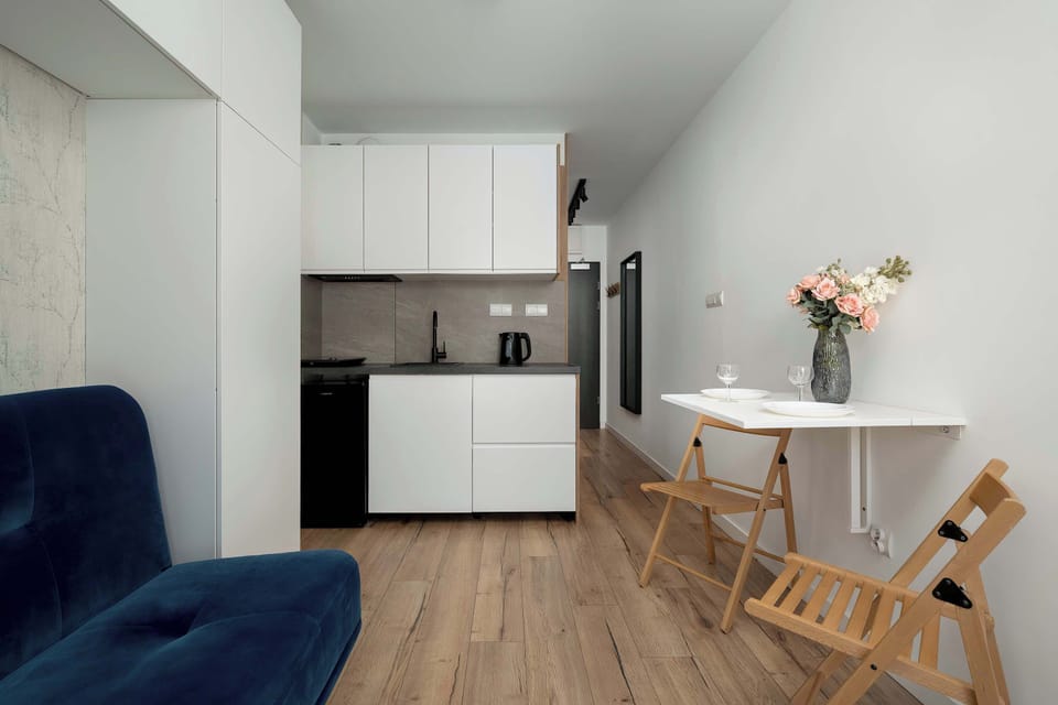 An open-plan room that combines a sitting area and a kitchenette. A blue sofa sits near a small table and chairs, while white cabinets and a counter are visible in the kitchen area. The space has light wood floors and a minimalist design.