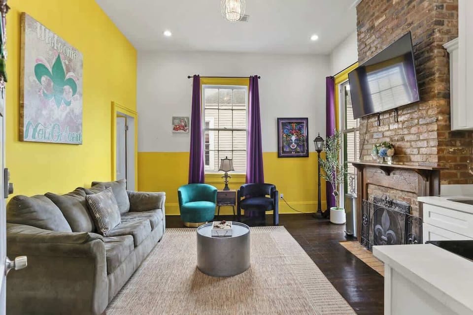 Vibrant living area with comfortable seating, exposed brick fireplace, and Smart TV. High ceilings, bold colors, and large windows create a bright, welcoming space to relax after exploring New Orleans.