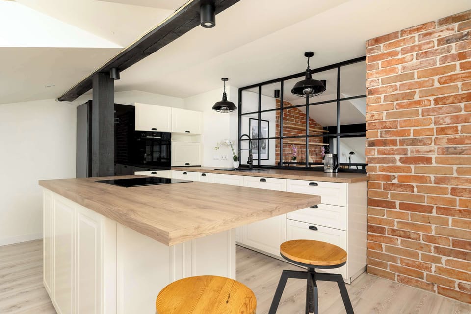 A rustic-modern kitchen island made of light wood serves as a focal point next to a textured red brick wall. Industrial black bar stools are tucked under the counter, blending warmth with an edgy design.