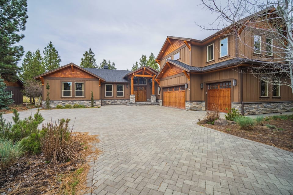 Stunning mountain lodge featuring rustic wood and stone architecture with spacious paver driveway and mature forest surroundings.