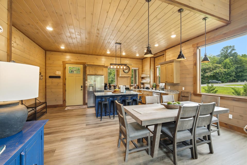 Gather around this spacious dining table or the inviting kitchen island, where rustic wood meets modern convenience. Large windows frame serene green views, filling your space with natural light and warmth.