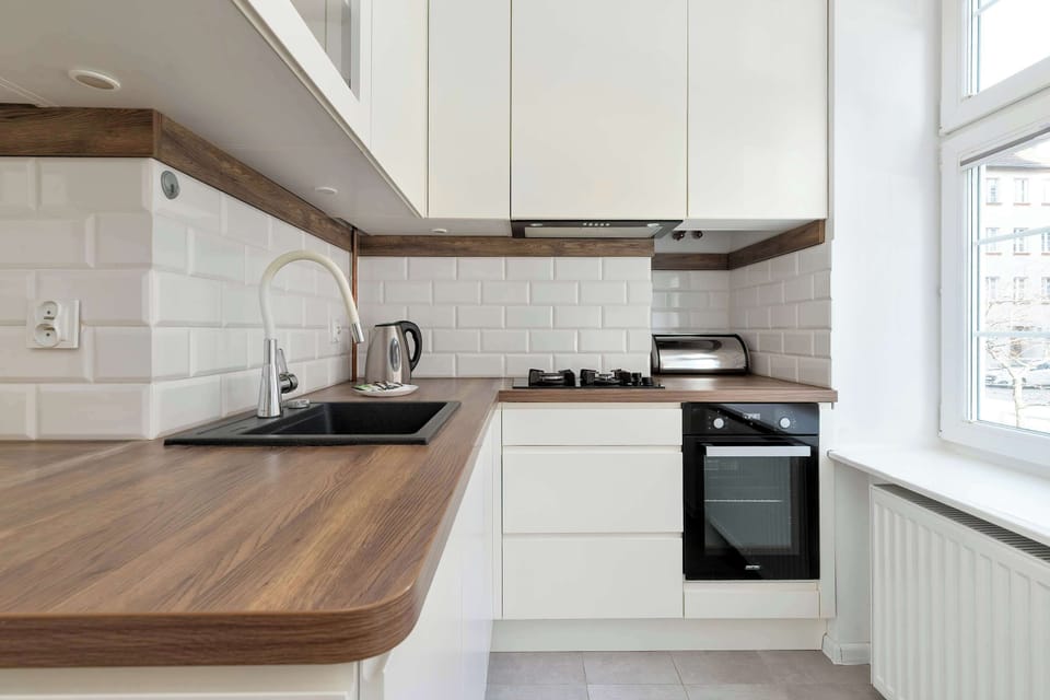 A compact L-shaped kitchen corner shows a white cabinet, a built-in oven, wood-grain countertops, and a window next to a radiator.