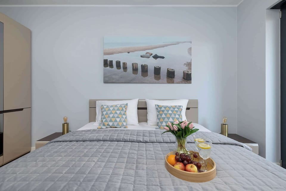 A comfortable bed arrangement highlights the grey bedding and a large window that lets in soft, natural light. A framed photograph of a pier hangs above the headboard, reinforcing the coastal theme.