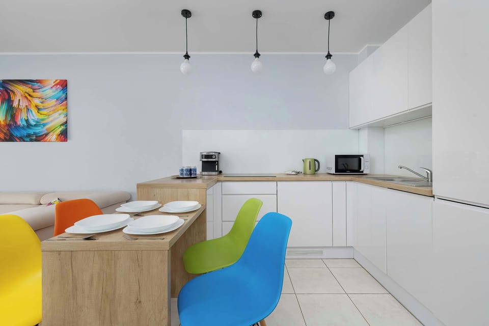 A modern kitchen and dining area includes sleek white cabinets and a wooden breakfast bar. The space is energized by vibrant dining chairs in yellow, orange, and blue.