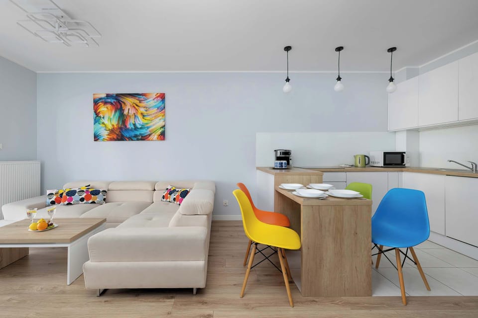 A wide-angle shot of the living area captures the harmony between the white kitchen and the vibrant seating zone. The light wood flooring flows throughout, unifying the different areas of the apartment.