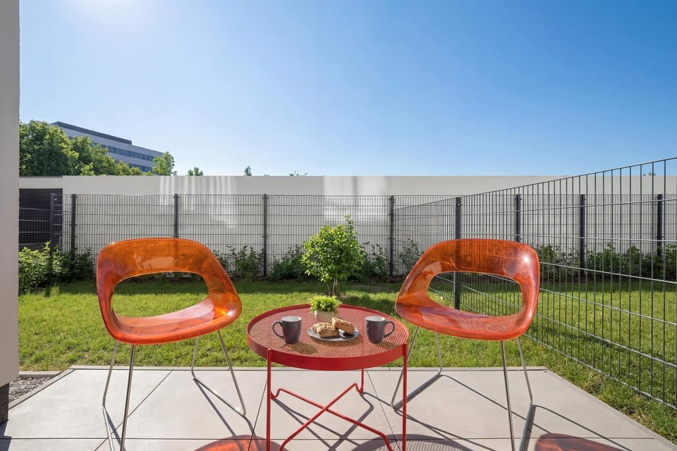The private garden offers a vibrant outdoor escape with two bright orange designer chairs and a matching small round table. A green lawn and a modern fence complete the space, providing a sense of seclusion.