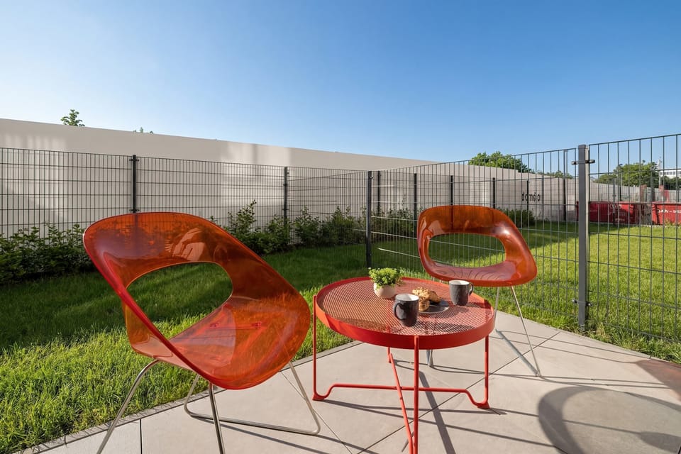 The garden area is seen from a low angle, highlighting the bright orange patio furniture on the stone tiles. The lush green grass and clear blue sky create an inviting atmosphere for outdoor relaxation.