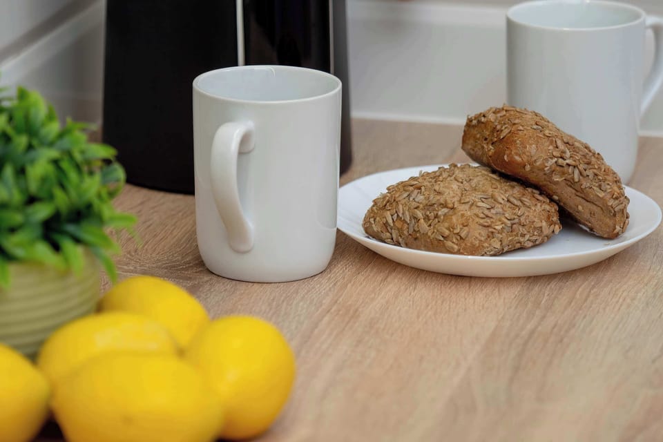 A minimalist-style kitchen area features a light-colored wooden countertop with a bowl of yellow lemons, a white mug, a wooden plate with food, and a small plant.