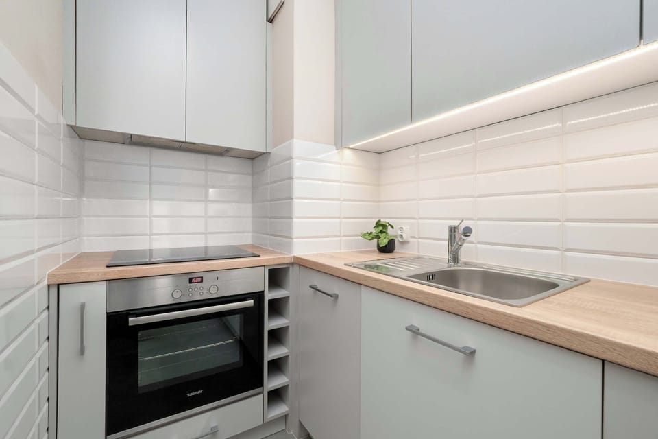 A minimalist-style kitchen area is equipped with sleek white cabinets, stainless steel appliances, a modern backsplash, and ambient overhead lighting, all set above a light-colored wooden countertop and white tiled backsplash.