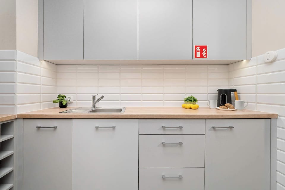 A minimalist-style kitchen area features light-colored wooden countertops, white subway tiles, sleek white cabinets, and stainless steel appliances, with a red sign on the cabinet door.