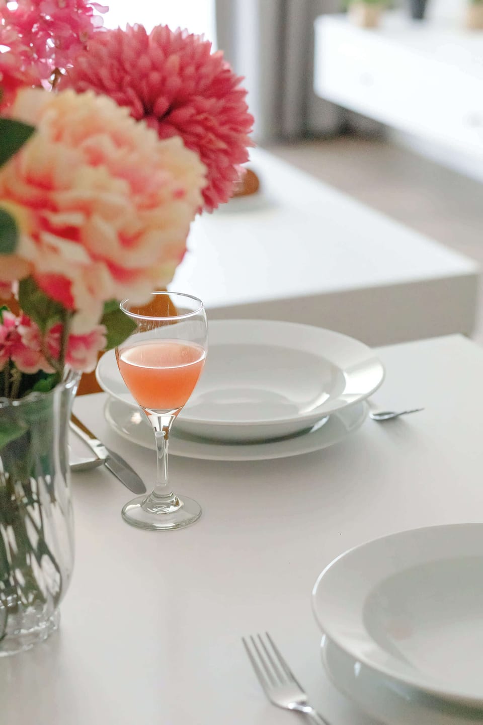 A vertical shot focuses on a glass of juice and white dinnerware on a dining table. Soft pink and red flowers in the background provide a lovely, blurred pop of color.