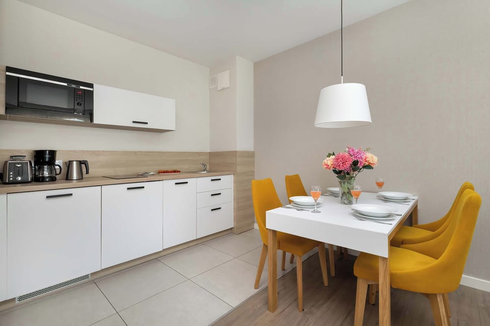 The kitchenette area consists of minimalist white cabinetry and a light wood backsplash. A small dining table with four yellow upholstered chairs provides a cheerful spot for meals under a pendant light.