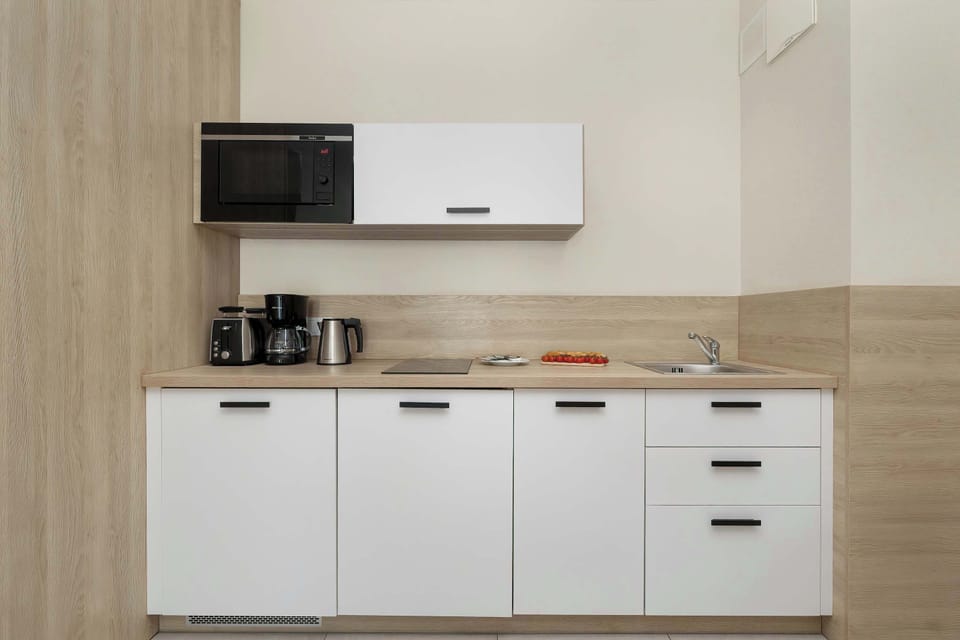 The kitchenette wall is showcased, featuring a built-in microwave and sleek, handle-less white cabinets. The wooden-textured backsplash adds warmth to the clean, white culinary space.