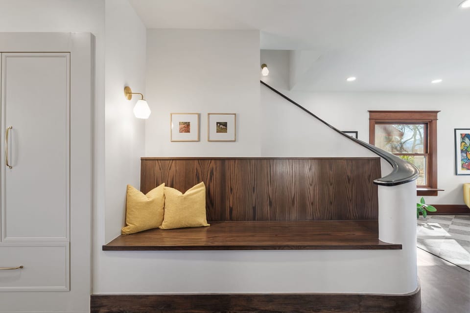 Relax on your custom walnut bench with cozy pillows, perfectly positioned to catch natural light from the beautiful wood-framed window.