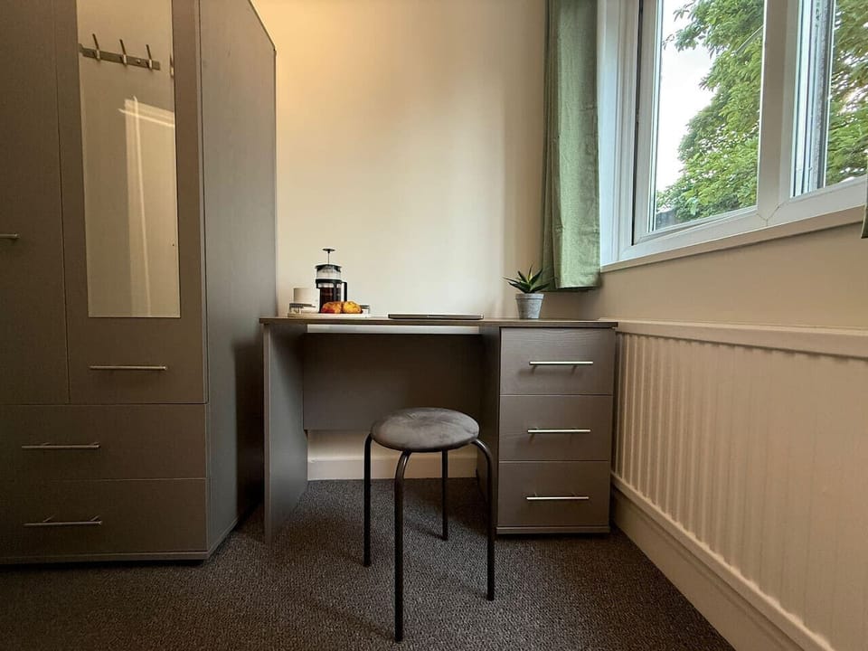 A bedroom corner showing a grey wardrobe, a desk with a stool, a radiator, and a window providing natural light — suitable for working from the room.