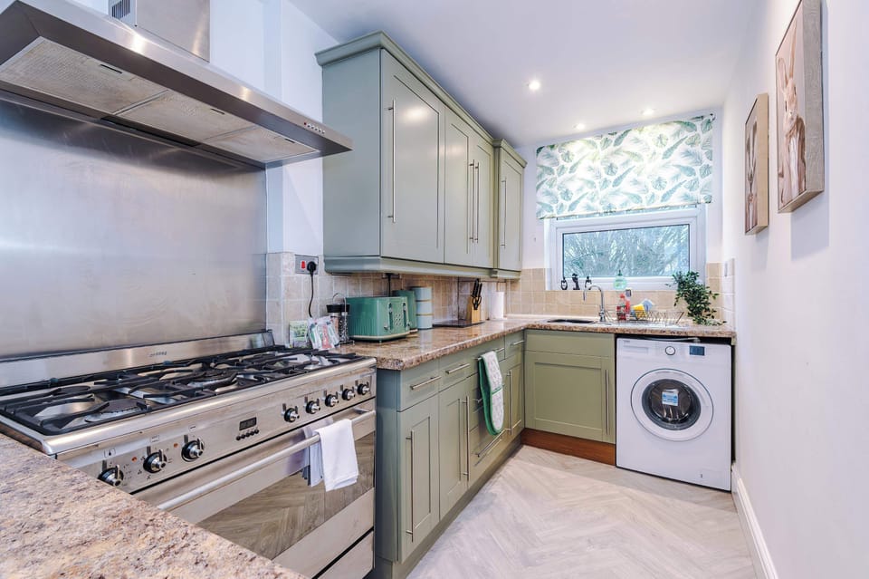 Stylish kitchen with muted green cabinets, granite countertops, and stainless steel appliances. Includes a large gas stove, washing machine, and window with leafy curtains—bright, functional space ideal for cooking and laundry convenience during your
