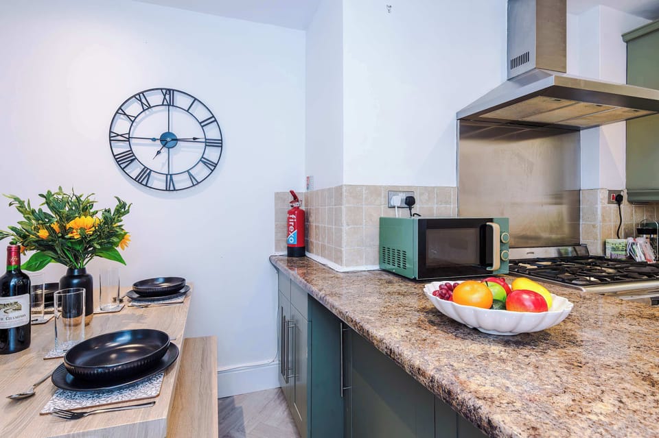 Stylish kitchen with muted green cabinets, granite countertops, and stainless steel appliances. Includes a large gas stove, washing machine, and window with leafy curtains—bright, functional space ideal for cooking and laundry convenience during your