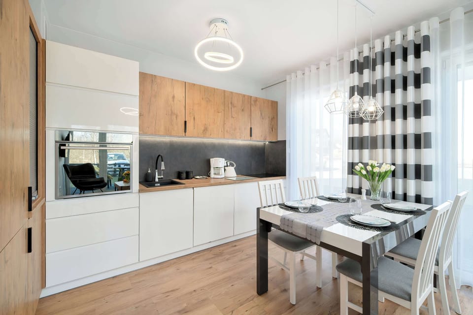 An open kitchenette and dining space with wooden cabinets and built-in appliances. A dining table stands in front of the kitchenette area. The design combines white walls with warm wooden textures.