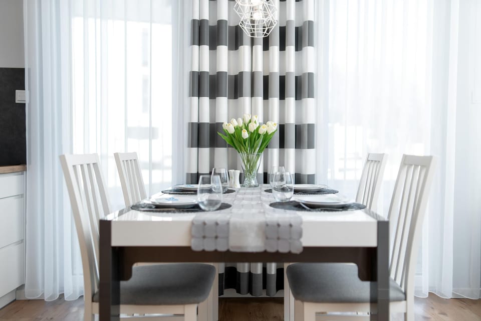 A dining area placed next to large windows with light curtains. A wooden table is prepared with plates and glasses. The space feels bright and welcoming thanks to natural light.