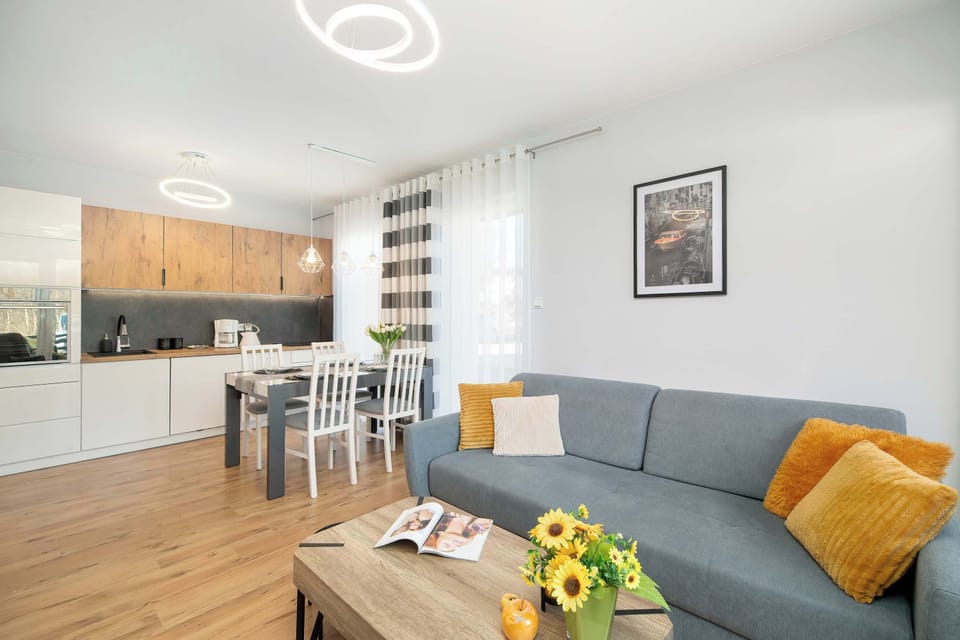 A modern living area with a gray sofa decorated with yellow accent pillows. A wooden coffee table with decorative items sits in front of the sofa. The open layout connects the living space with the kitchenette in the background.