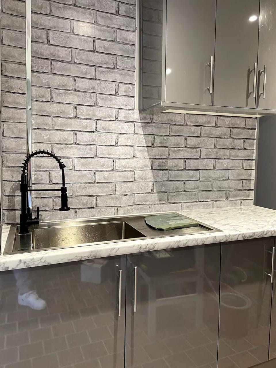 Kitchen worktop detail showing a black mixer tap, stainless steel sink, and brick-effect tiled splashback.