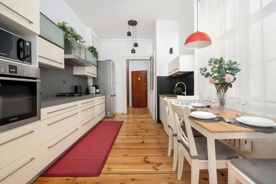 The kitchen is shown in full length, featuring a built-in oven, microwave, and a dining table at the far end. Potted plants on top of the upper cabinets bring a touch of greenery to the space.