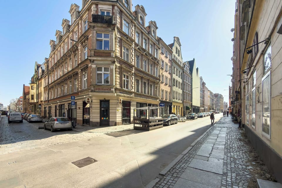 The exterior shot shows a beautiful, historic corner tenement house with ornate architectural details under a clear blue sky. The building is situated on a clean, cobblestone-adjacent street in an urban setting.