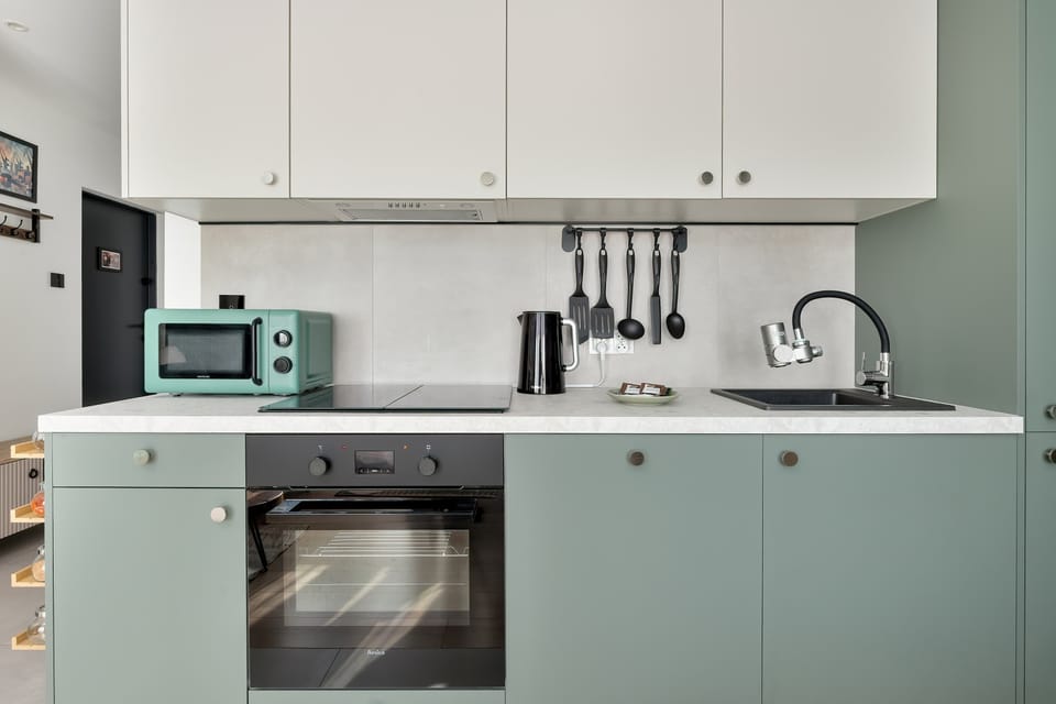 A compact kitchenette workspace with green cabinets, a sink, and a stovetop. White cabinetry above keeps the area bright and uncluttered. Everyday appliances are neatly integrated into the design.