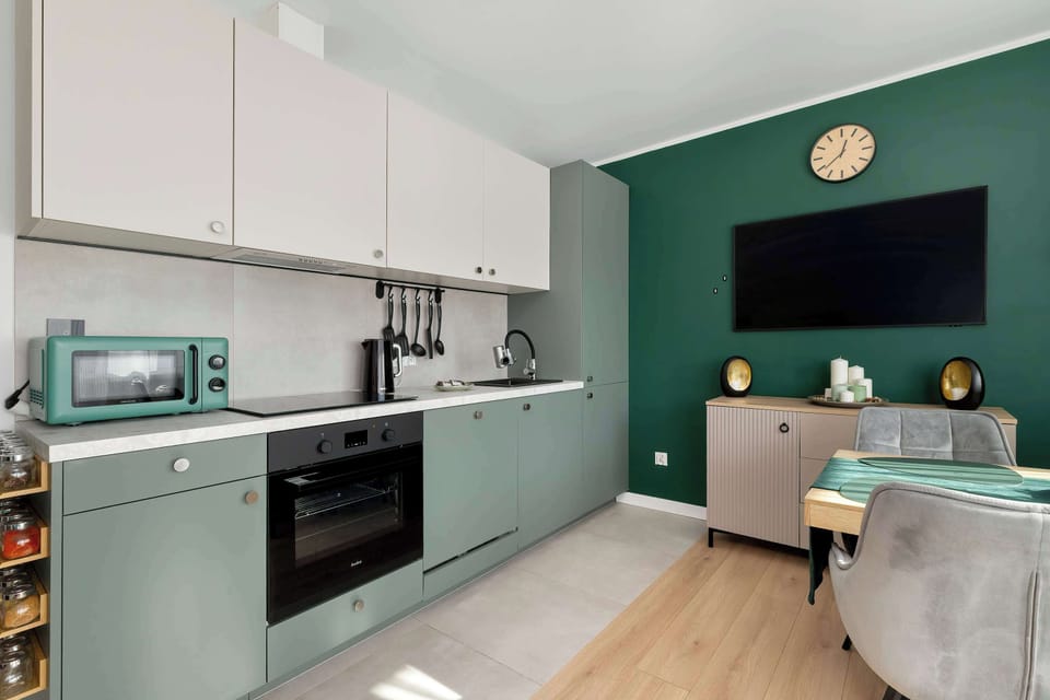 A long kitchenette view with cabinets, countertops, and built-in appliances along one wall. The green cabinetry contrasts with the white walls and light wood flooring. The layout appears practical for cooking and dining.