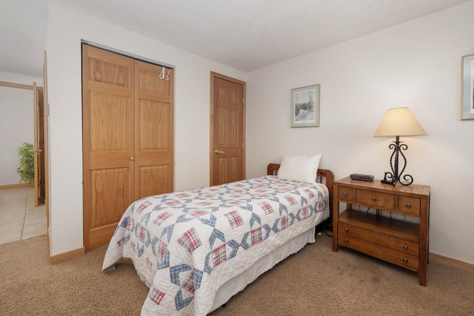 Your cozy bedroom features a comfortable twin bed with charming quilt, warm wood furnishings, and soft lighting for peaceful rest.