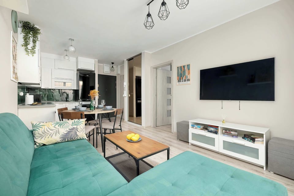 A modern, well-lit living room from an apartment includes a green L-shaped sofa on the left, a coffee table with stools, and a dining table with chairs, all on light wood flooring. 