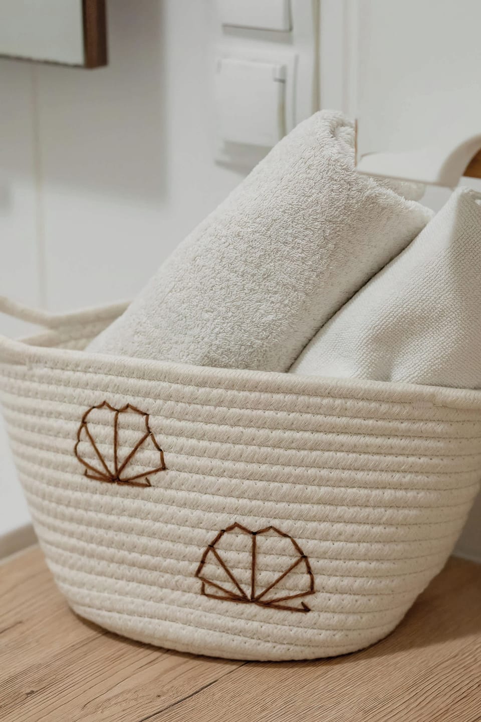 A cozy-looking, woven straw-like tote bag sits filled with rolled light-colored blankets. The bag features a repeating simple white sunburst design and is resting in front of a modern radiator and a white wall with a vertical pipe.