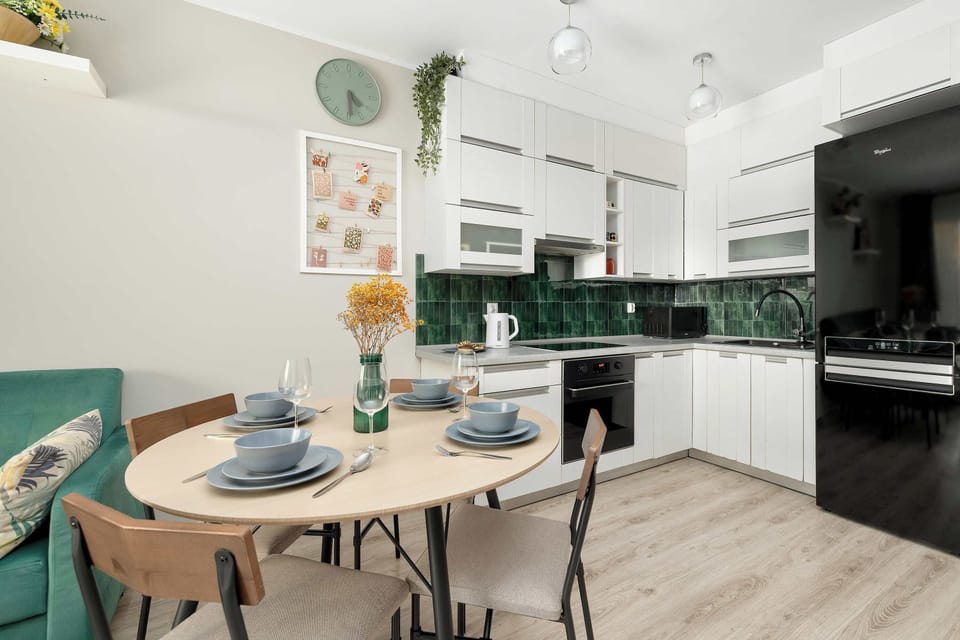 A round, light wood dining table set for five with grey plates and glassware in a kitchen-dining area. It has light wood flooring and is surrounded by five simple chairs with curved light wood seats.