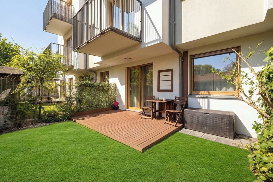 A well-maintained, private terrace includes a large lawn, a paved area, and comfortable outdoor wooden furniture with dark cushions. The low-rise, modern apartment building with balconies is visible in the background against a clear blue sky.