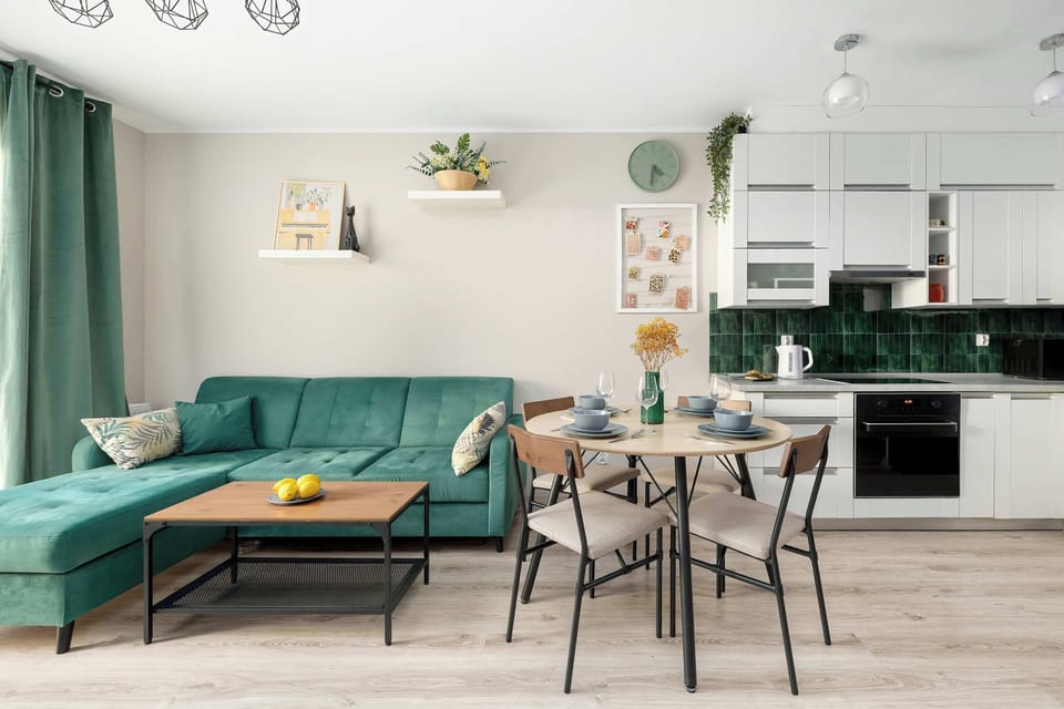 A bright, open-plan living and dining area in an apartment includes a green sofa on the left, a coffee table with two stools, and a dining table with chairs. At the back is a corner kitchen with white cabinets and a green backsplash.