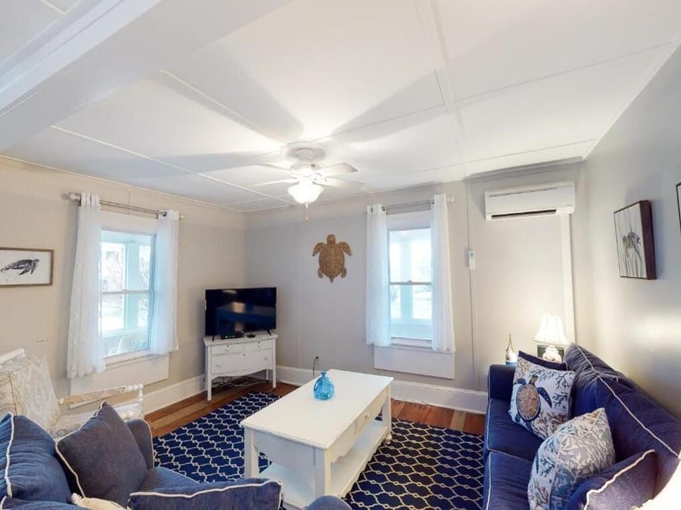The cozy living room features a blue sectional sofa, a coffee table, and a TV, illuminated by natural light from the dual windows with white curtains.