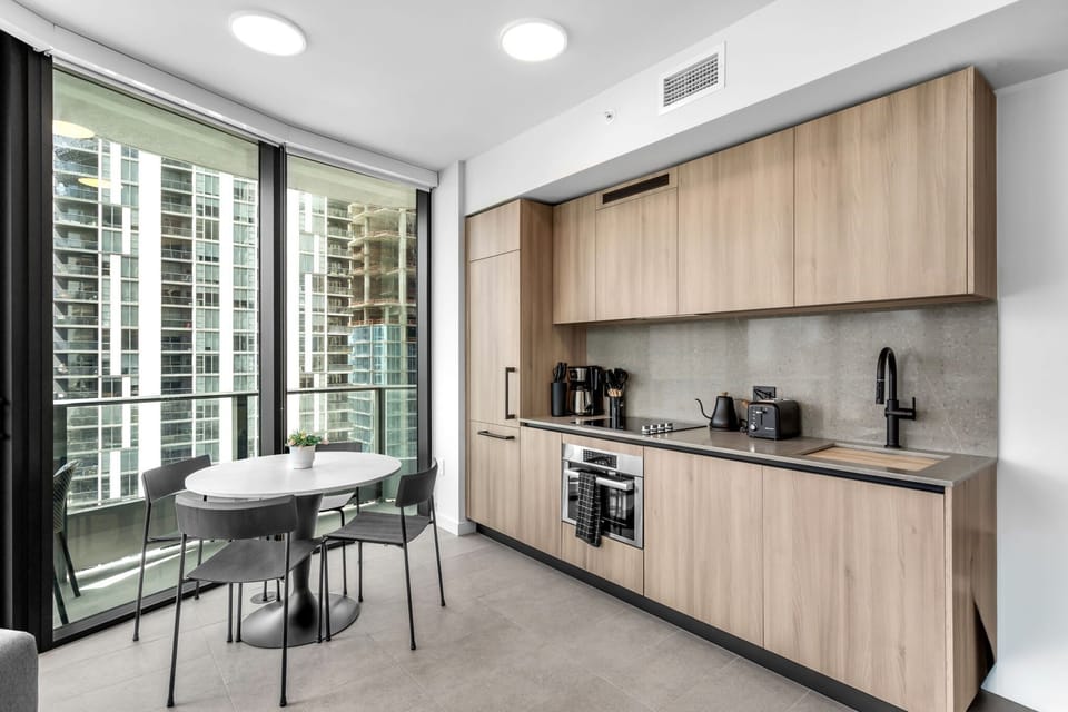 Cook and dine with a view! This sleek modern kitchen features light wood cabinetry and opens to a sun-drenched dining area with floor-to-ceiling windows. A perfect spot for home-cooked meals with a stunning city backdrop.