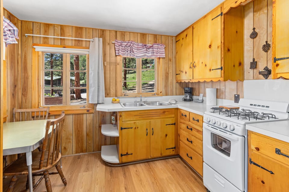 Wood-paneled kitchen with honey pine cabinets, white countertops, a double sink, gas stove, and a small dining nook by windows.