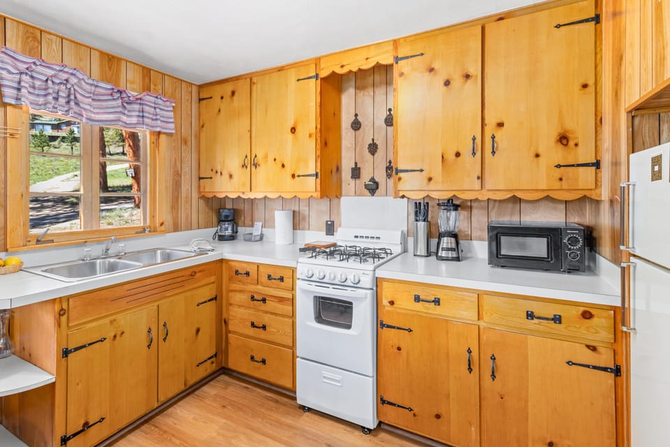 Kitchen with honey pine cabinets, white gas stove, double sink beneath striped valance window, and white countertops.