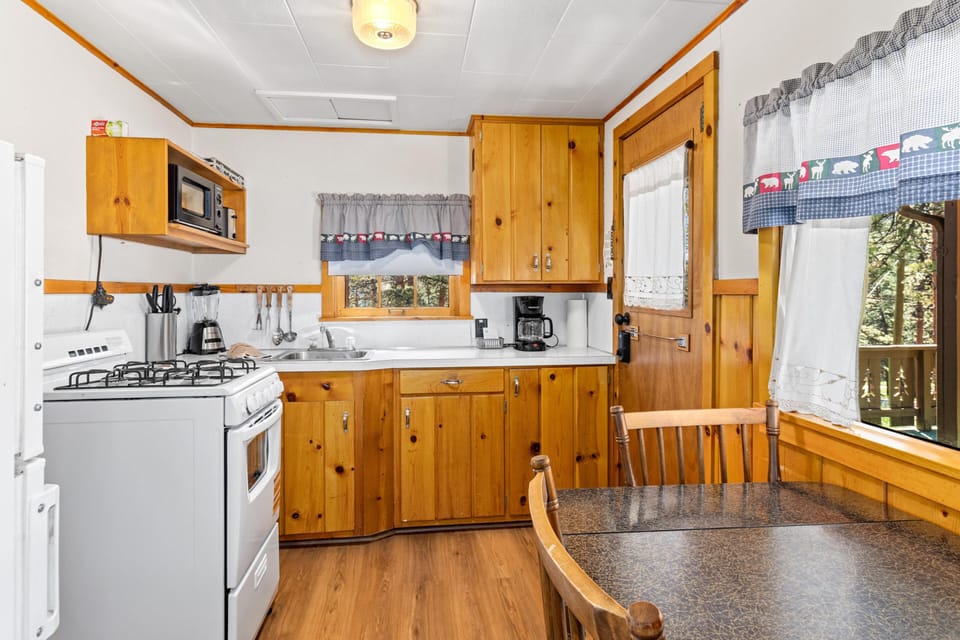 Wood-paneled kitchen with white countertops, gas stove, coffee maker, and window views of pine trees.