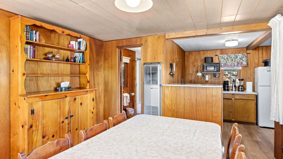 Pine-paneled dining area with wood chairs and a floral tablecloth, adjacent to a compact kitchen with white appliances.
