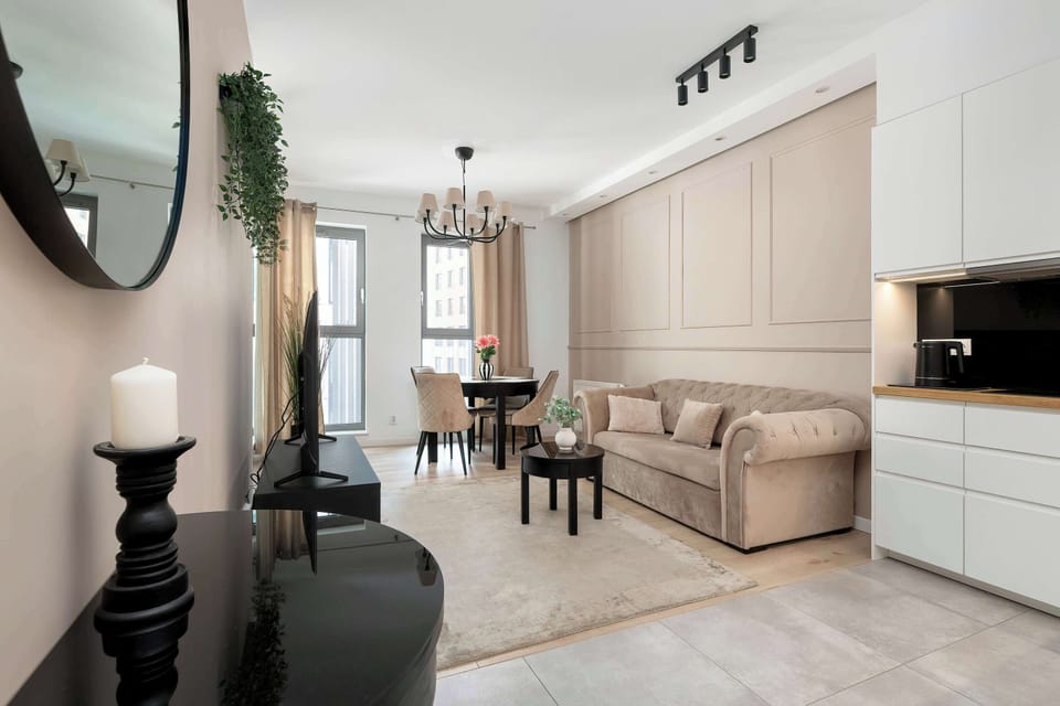 A bright living area connected to a kitchenette in a modern studio apartment. A beige sofa, dining table, and kitchen cabinets create a comfortable open-plan layout. Light wooden floors and neutral colors give the space a clean and elegant feel.