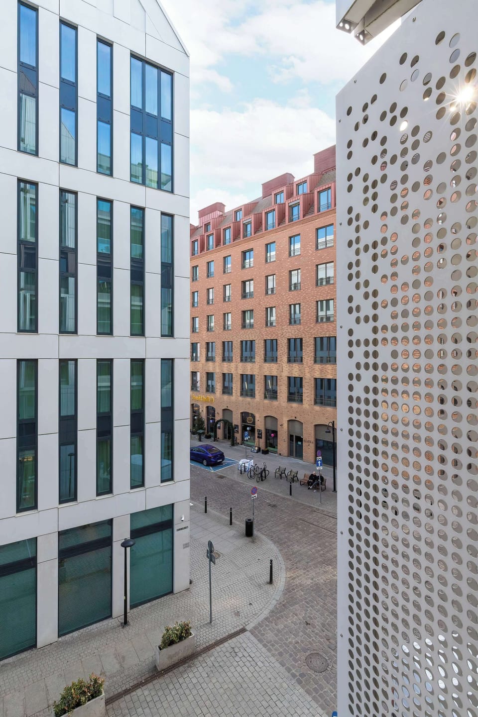 A narrow view between modern city buildings with tall glass and concrete facades. The area looks like a quiet urban passage or courtyard. The architecture reflects a contemporary city environment.