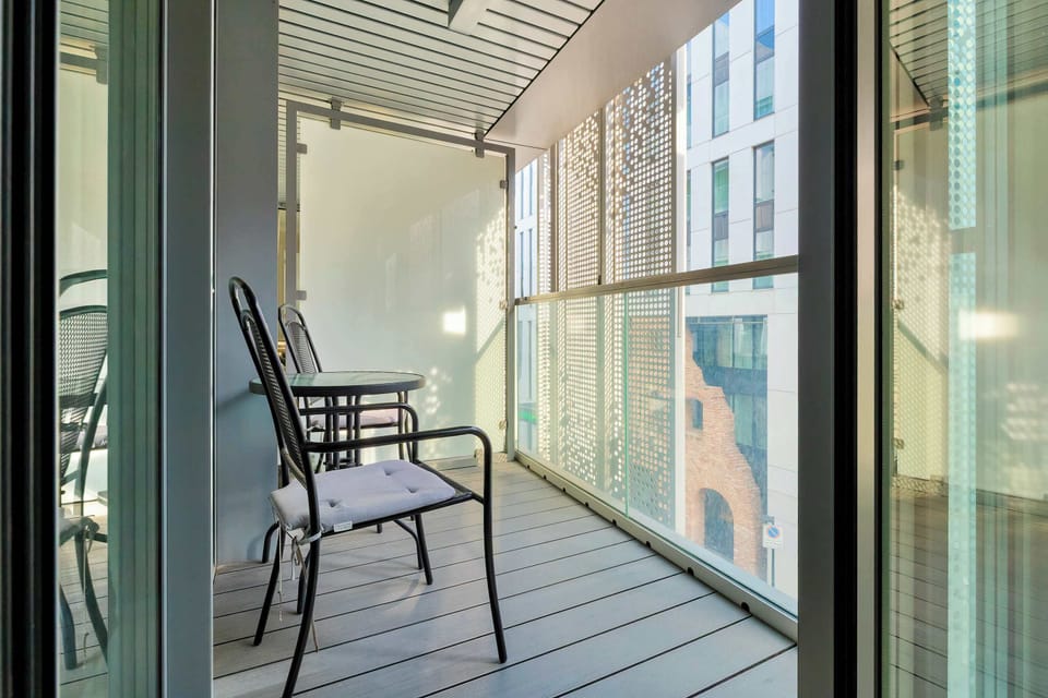A private balcony with glass railings and a view of nearby buildings. A small chair and table provide a comfortable outdoor seating area. It is a perfect place to relax and enjoy fresh air.