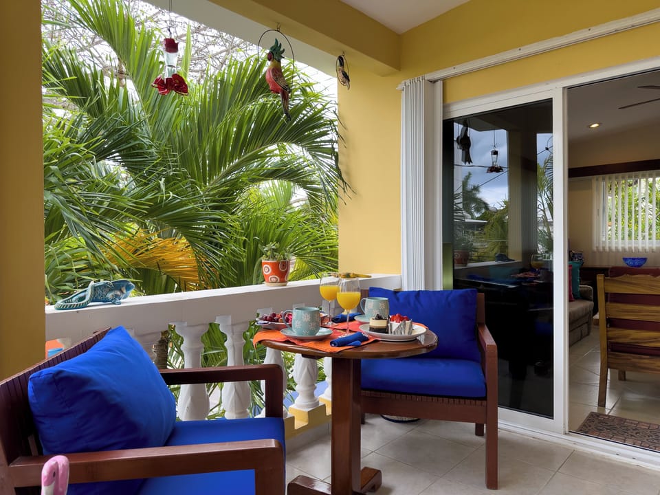 Abbey del Sol Apt 6 – Balcony Breakfast Setup - Private balcony set for breakfast with cushioned seating and a small table, surrounded by palms and tropical greenery.