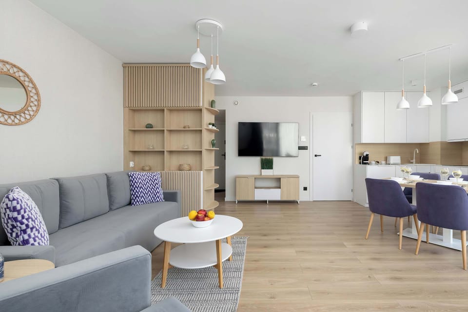 A stylish dining table is surrounded by elegant chairs with purple upholstery, positioned near a large window. The wooden shelving unit behind the table separates the dining zone from the rest of the room.