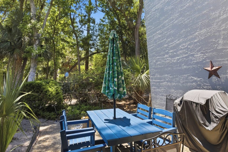 Azure blue furniture pops against the textured white stone wall of this shaded patio. Tall oaks and tropical fronds frame your private lunch spot, where the afternoon glow creates a tranquil setting for outdoor dining.