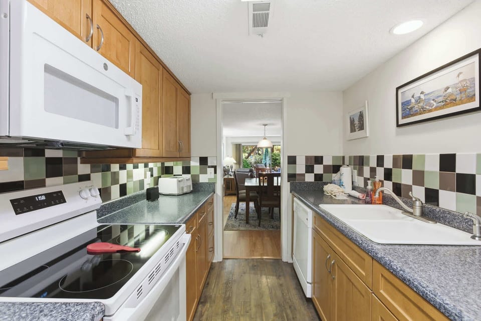 Chequered tiles in earthy tones bring a playful character to this efficient kitchen, where warm wood cabinetry meets speckled stone counters. Bright overhead lighting illuminates the path toward a cozy dining area and a view of the garden beyond.