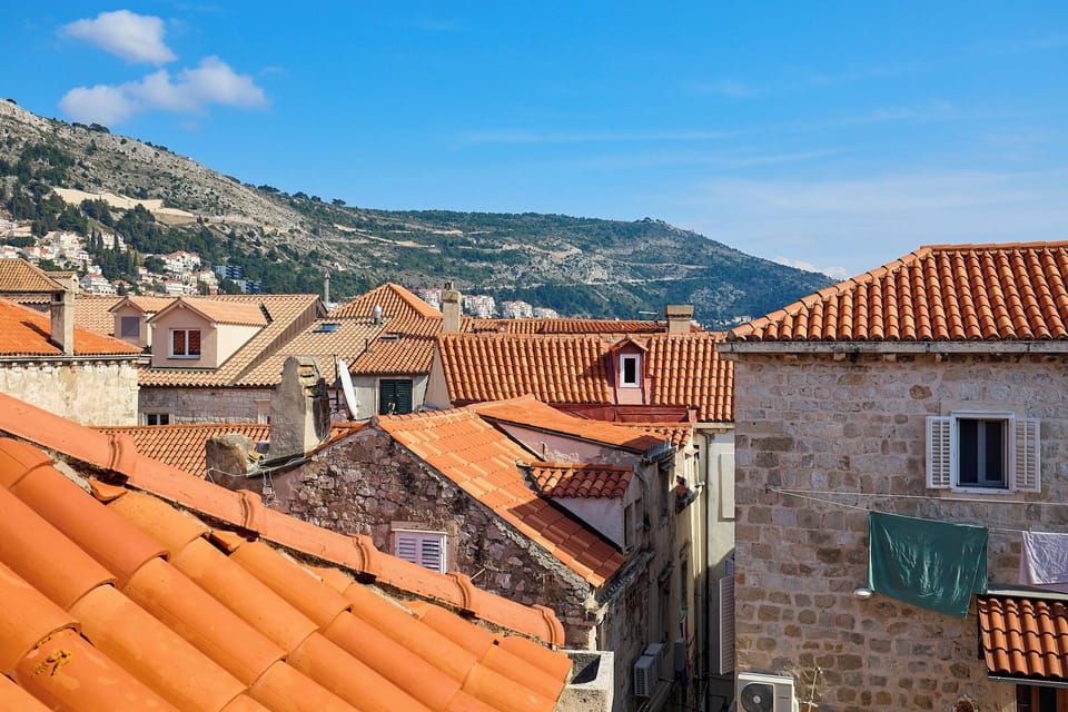 Gaze across the iconic terracotta rooftops of Dubrovnik's Old Town, with a stunning backdrop of the verdant hills under a clear blue sky.