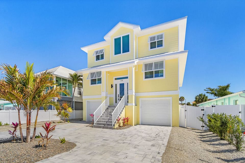 Stunning yellow beach house featuring a spacious driveway, dual garages, and a beautiful tropical entrance.
